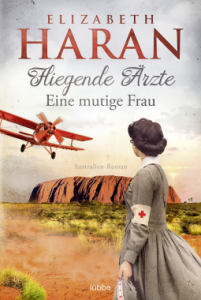Cover von "Fliegende Ärzte: Eine mutige Frau". Ein Frau steht mit dem Rücken zum Uluru in Australien und blickt zu einem Flugzeug am Himmel.